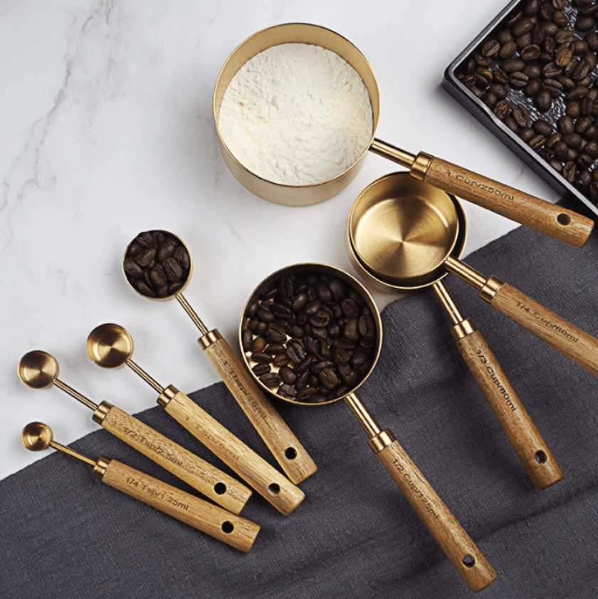 Set of brass measuring cups and spoons with wooden handles on a marble surface with coffee beans and flour.