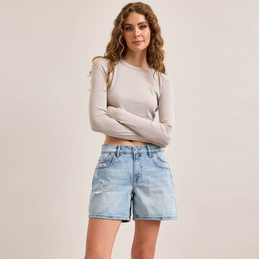 Woman wearing a beige long-sleeve top and light blue denim shorts on a plain background