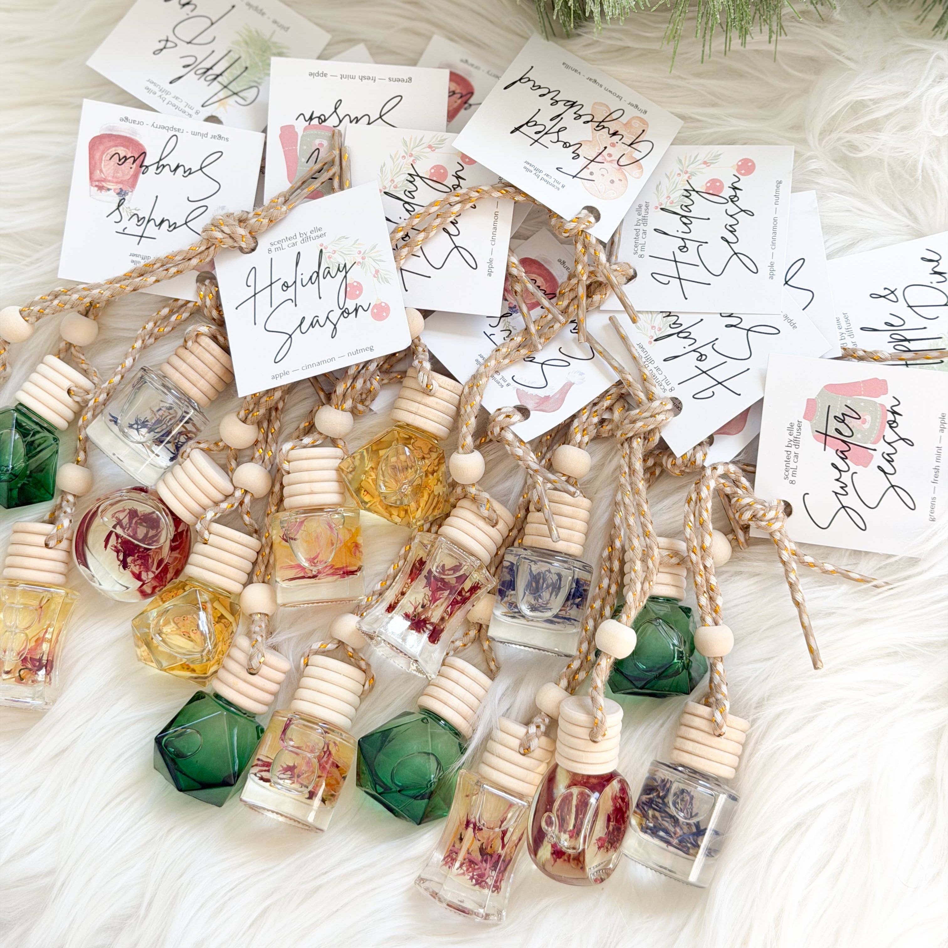 A styled flat-lay of small glass perfume bottles with wooden caps and dried botanicals inside, each attached to a handwritten gift tag and jute string. The bottles are arranged on a white faux-fur surface with green pine branches and a pinecone at the top.