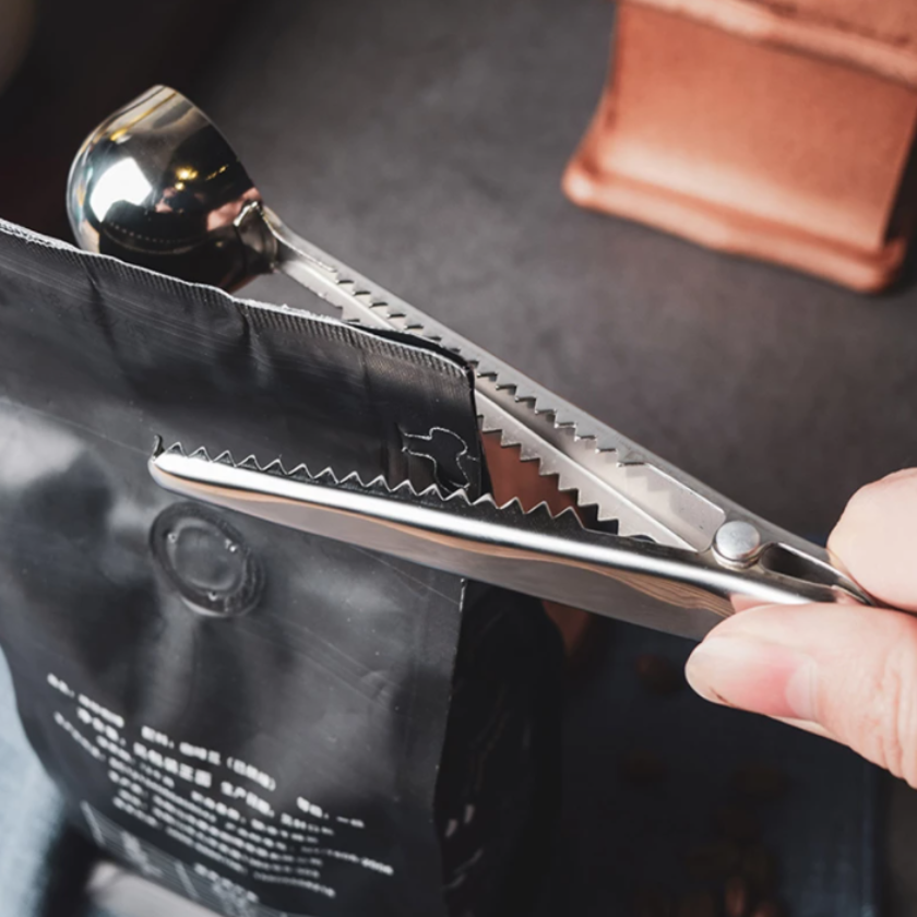 Metallic scissors cutting a black coffee bag with a brown leather item in the background.