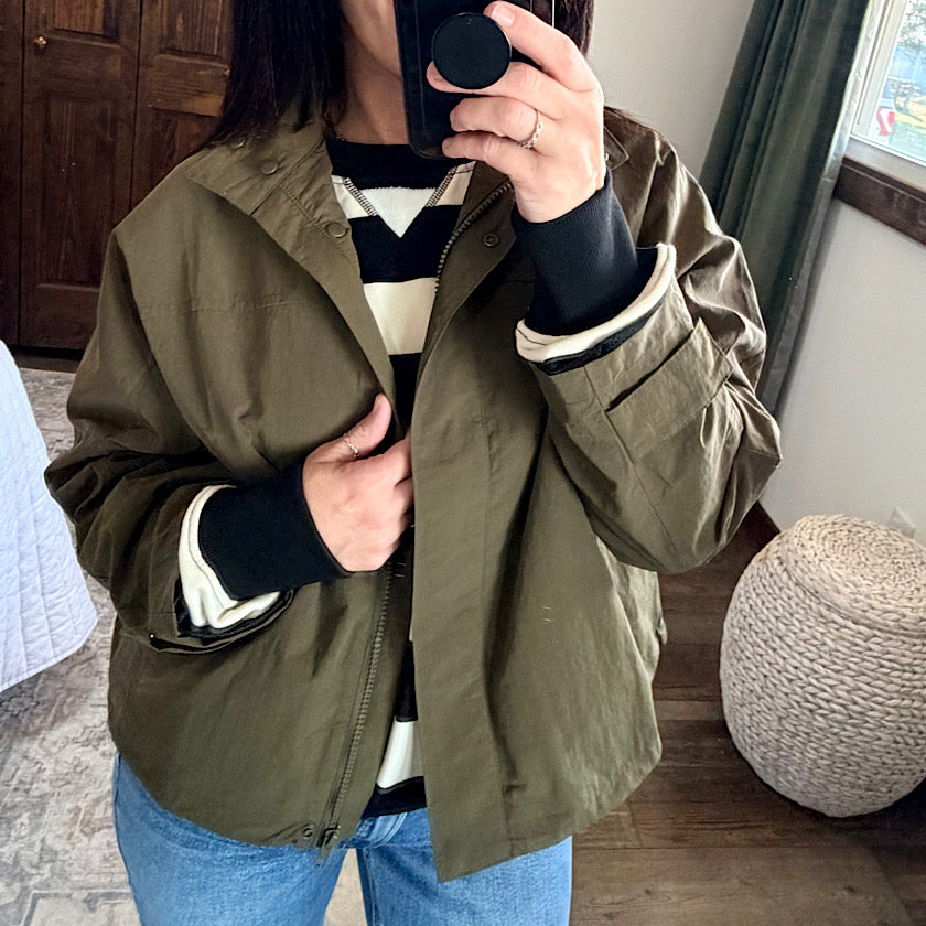 A person taking a mirror selfie wearing an olive green lightweight jacket over a black and white striped sweater, paired with blue jeans.