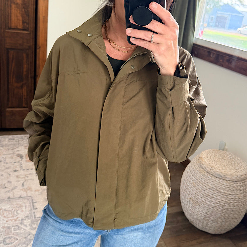 A person taking a mirror selfie wearing an olive green lightweight zip-up jacket over a black top and blue jeans in a home setting.