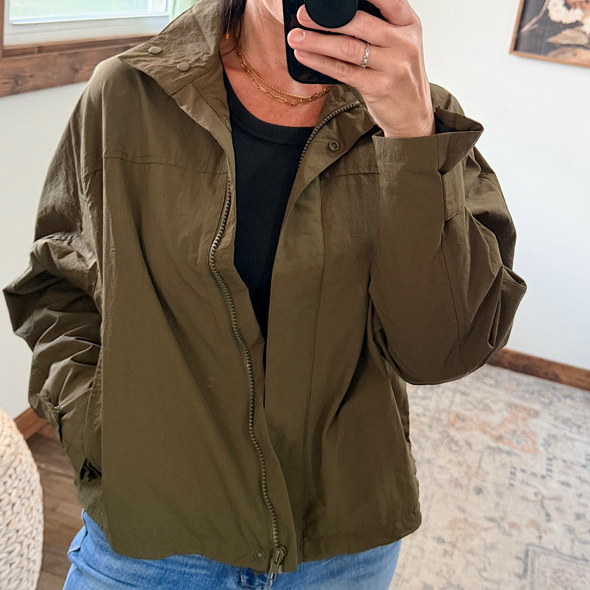 A person takes a mirror selfie wearing an olive green lightweight zip-up jacket over a black top and blue jeans in a home setting.