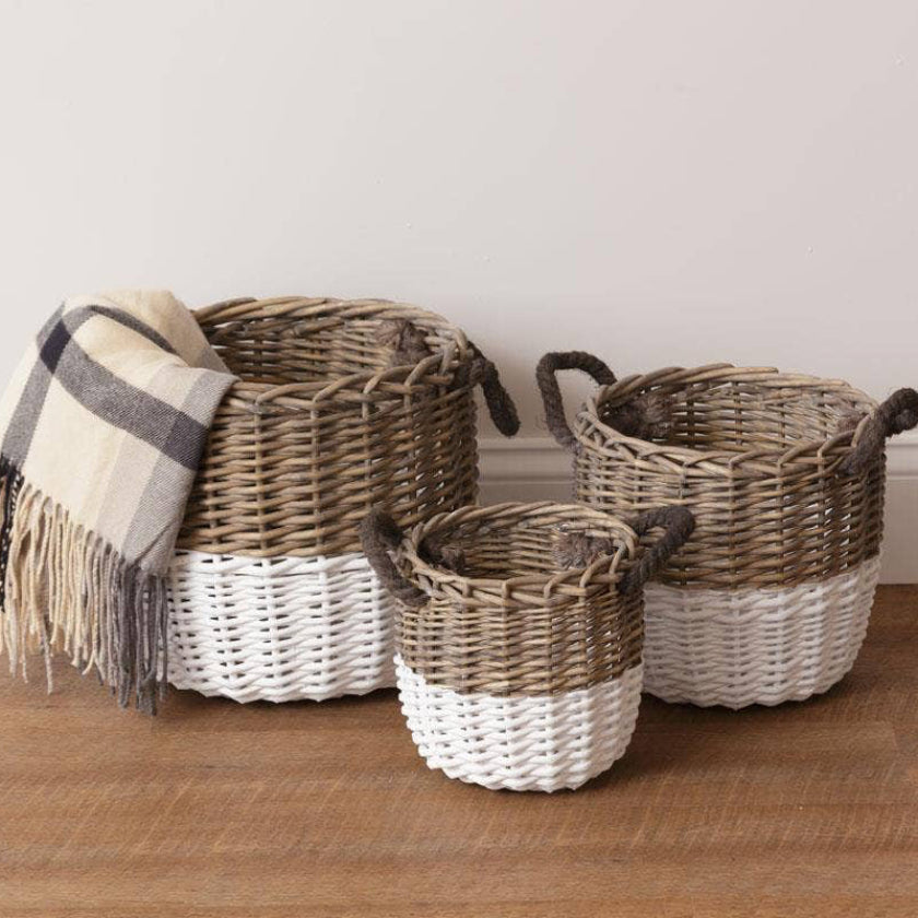 Three woven storage baskets in varying sizes with dark handles, featuring a two-tone design of natural brown wicker and white woven lower half. A beige and gray plaid throw blanket is draped over the largest basket, which is set on a wooden floor against a neutral beige wall.