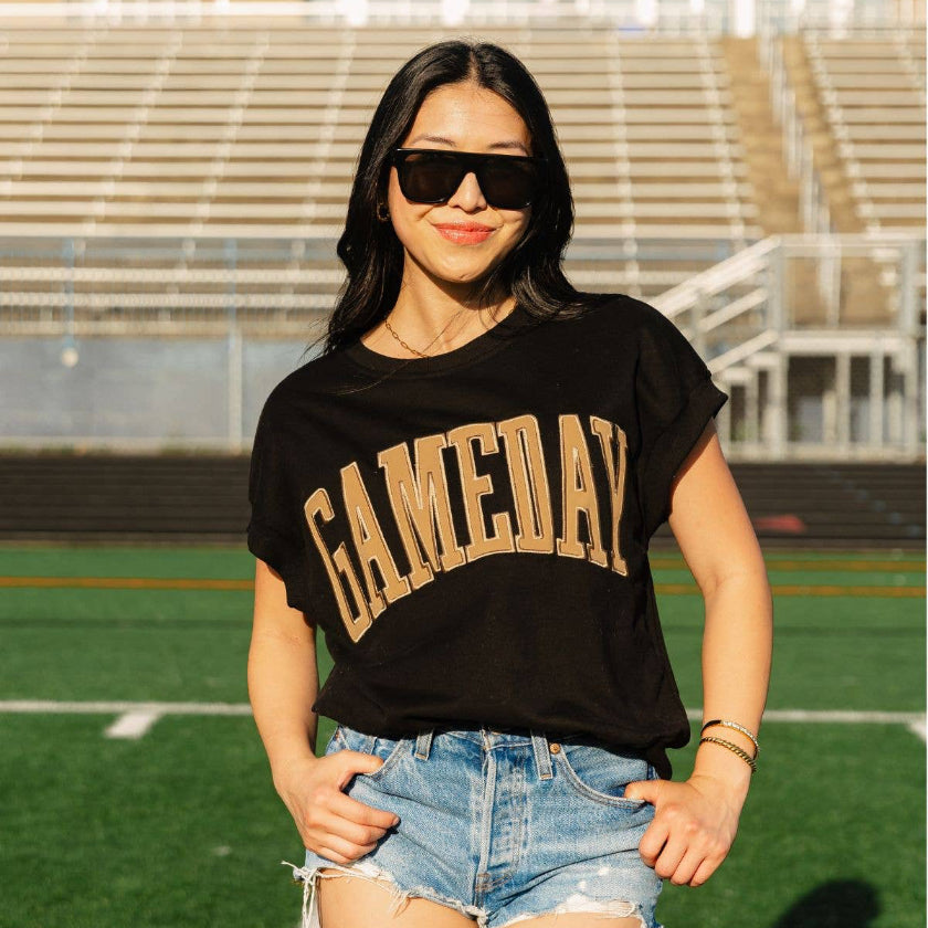 A woman posing on a football field wearing a black graphic t-shirt with 'GAMEDAY' printed in gold arched letters, paired with denim shorts and black sunglasses.