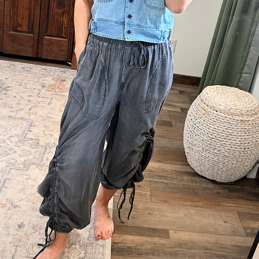 A person wearing loose dark gray drawstring pants with side ruching and gathered cuffs, paired with a light blue denim vest, standing on a patterned rug over a wood floor in an indoor room.