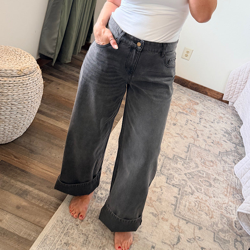 A person wearing dark gray wide-leg cuffed jeans and a white top, standing barefoot on a patterned area rug in a bedroom setting.
