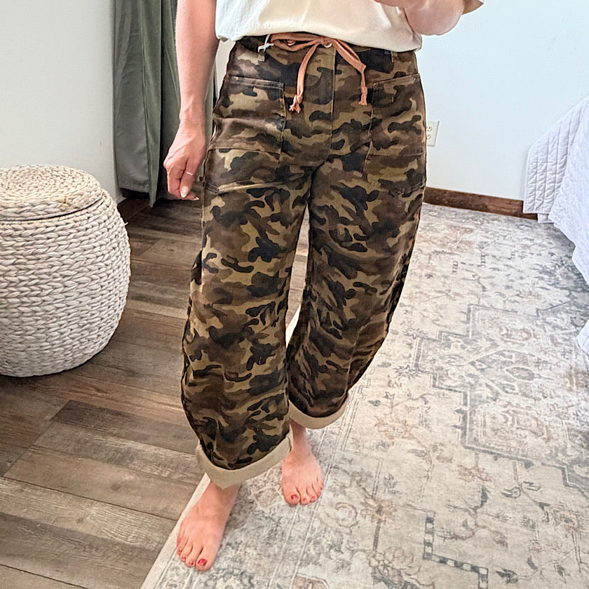 A person wearing loose-fit camouflage pants with a tan drawstring waist, standing barefoot on a patterned rug in a bedroom setting.