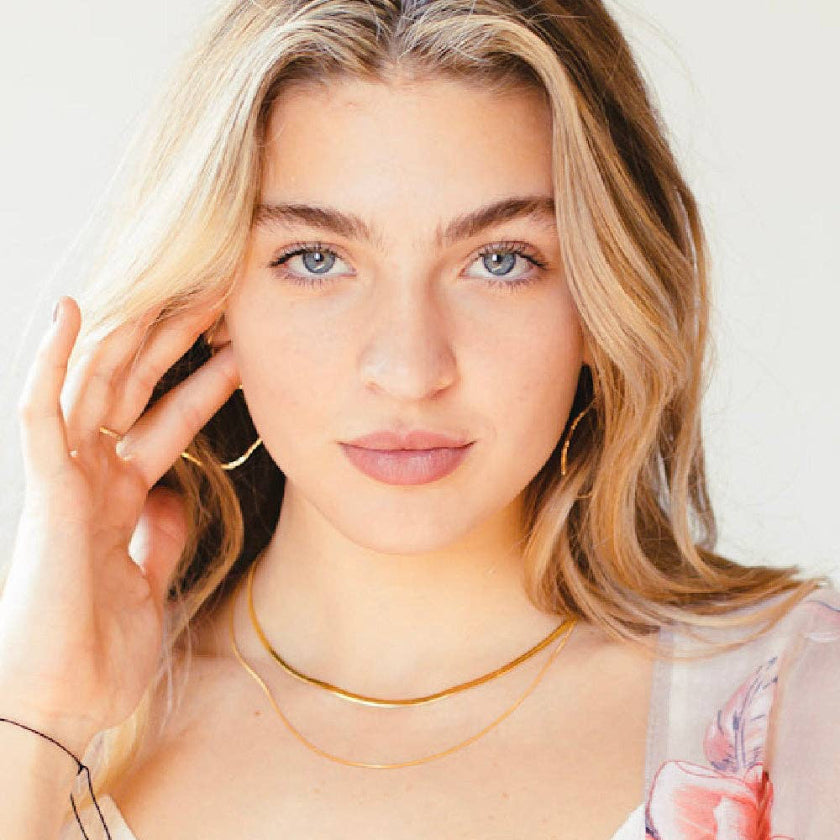 Woman with blonde hair and blue eyes wearing a gold necklace against a white background