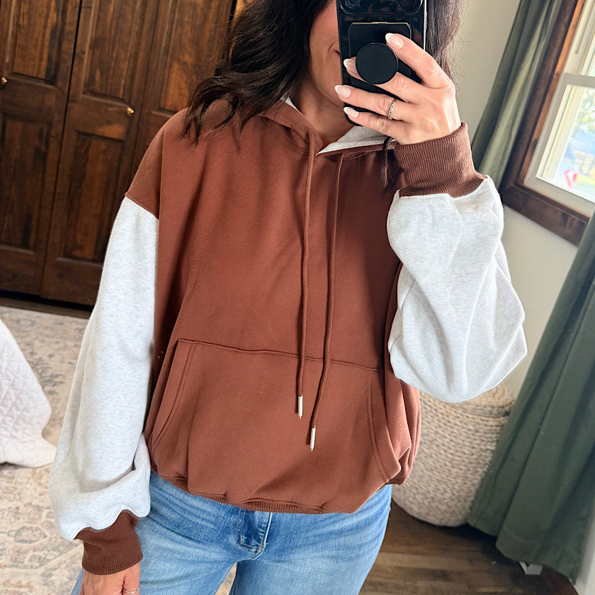 A person taking a mirror selfie wearing a color-blocked hoodie with a brown body and light gray sleeves, paired with blue jeans in a bedroom setting.