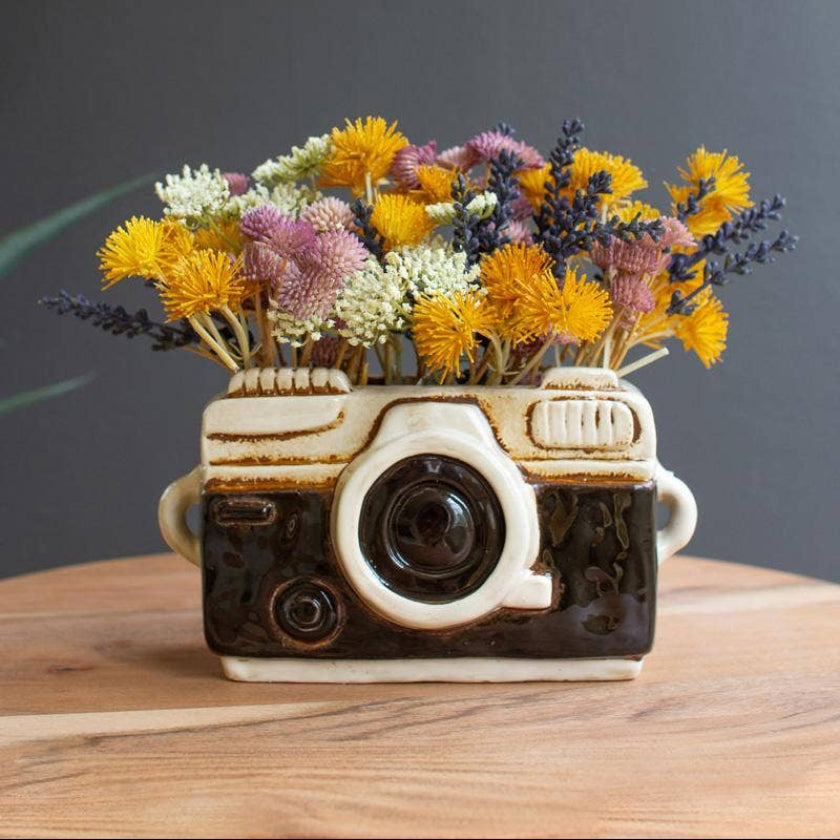 Ceramic camera-shaped vase with flowers on a wooden surface against a dark background