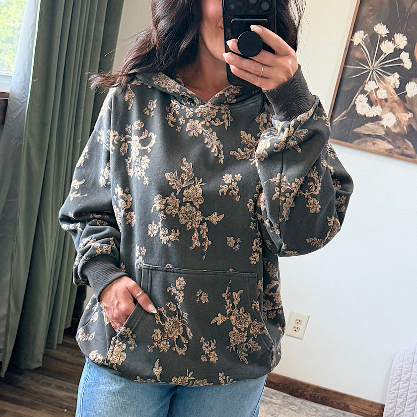 A woman takes a mirror selfie wearing a dark olive green hoodie with a beige floral pattern, paired with blue jeans in a home setting.
