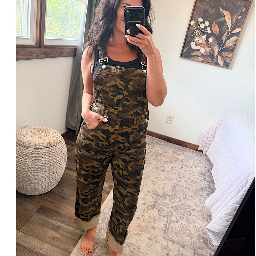 A woman takes a mirror selfie in a bedroom wearing camouflage print overall with a black tank top underneath. The room has a patterned rug, a woven ottoman, and a framed botanical painting on the wall.