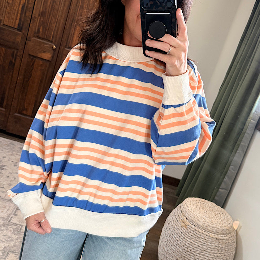 A person takes a mirror selfie wearing an oversized long-sleeve crewneck sweatshirt with horizontal stripes in blue, peach, and cream, paired with light blue jeans.