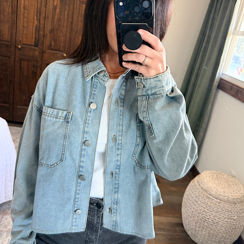 A person takes a mirror selfie wearing a light-wash blue denim button-up shirt over a white top and dark jeans, holding a black smartphone with a pop socket.