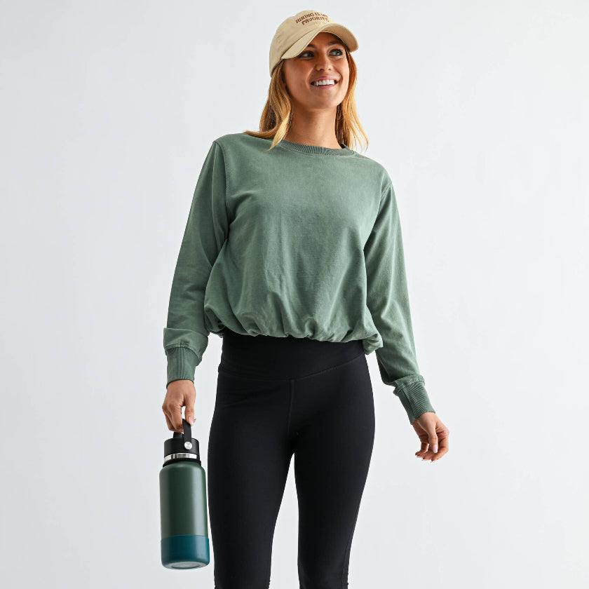 Woman wearing a green long-sleeve shirt and black pants, holding a water bottle on a white background