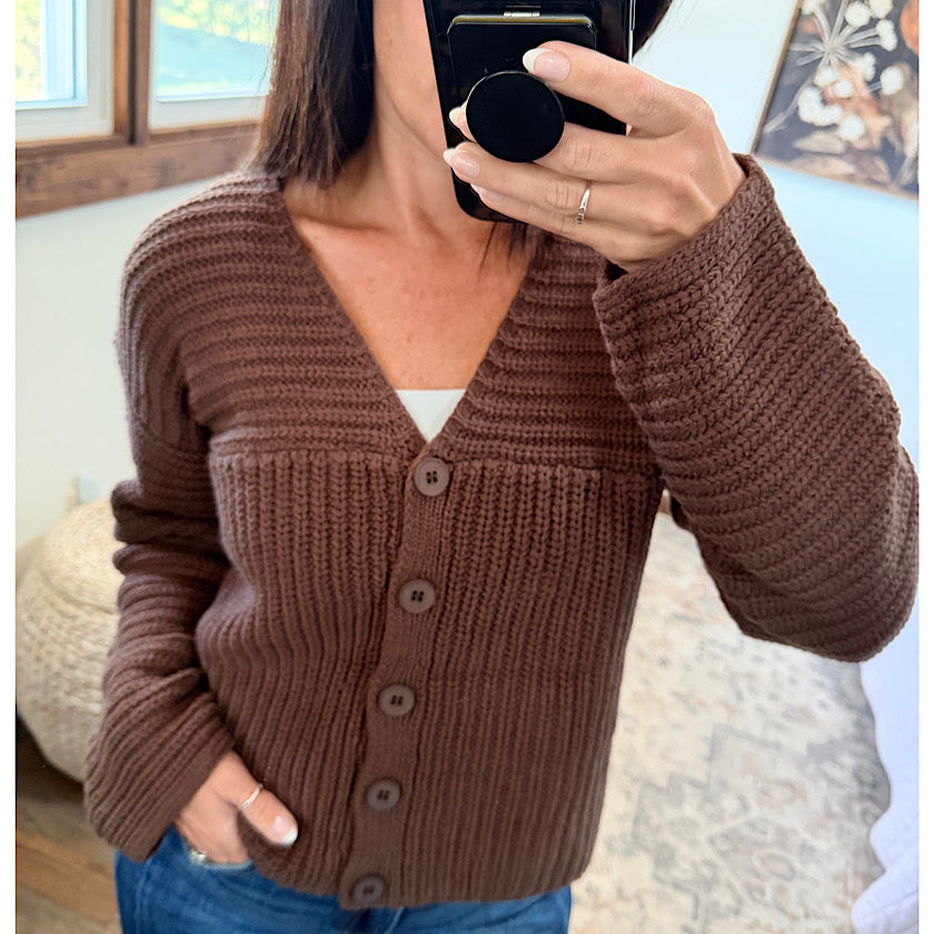 A woman takes a mirror selfie showing a brown chunky knit cardigan sweater with buttons, paired with blue jeans.