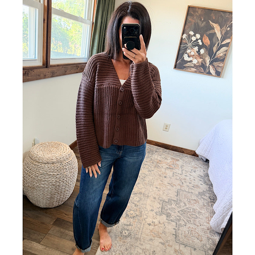 A woman takes a mirror selfie in a bedroom wearing a brown ribbed button-front cardigan over a white top and blue jeans.