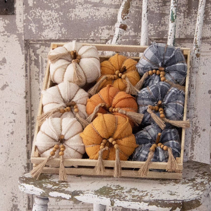 A wooden crate filled with decorative fabric pumpkins in cream, mustard yellow, orange, and blue plaid, arranged on a distressed white chair against a rustic weathered wall.