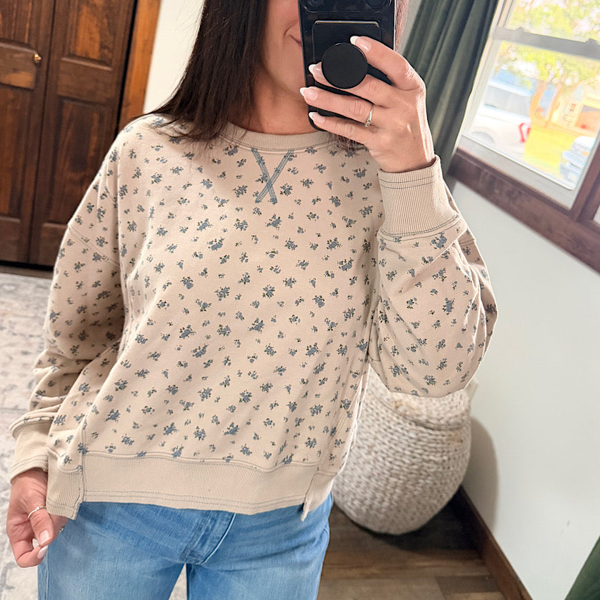 A woman takes a mirror selfie wearing a beige crewneck sweatshirt with a small gray floral pattern and ribbed cuffs, hem, and collar, paired with blue jeans in an indoor setting.