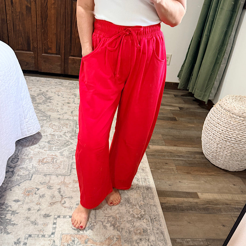 A person wearing bright red drawstring wide-leg pants, standing barefoot on a patterned rug in a bedroom setting.