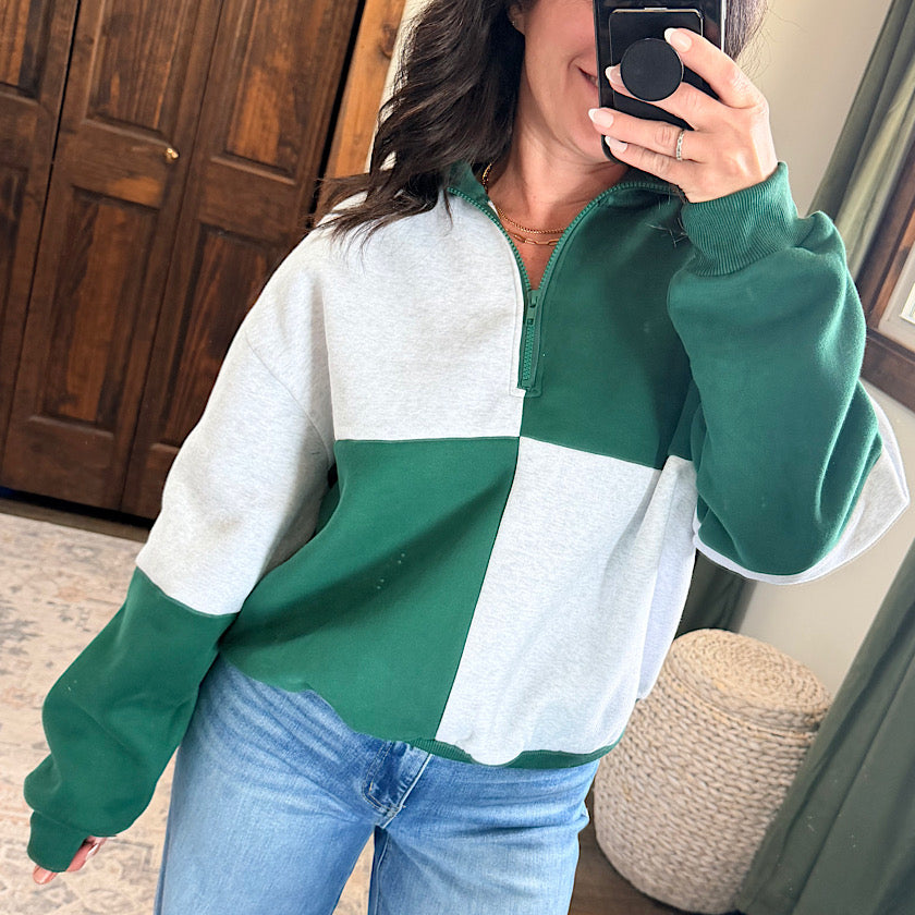 A woman takes a mirror selfie wearing a color-blocked quarter-zip sweatshirt with green and light gray blocks, paired with blue jeans in a home setting.