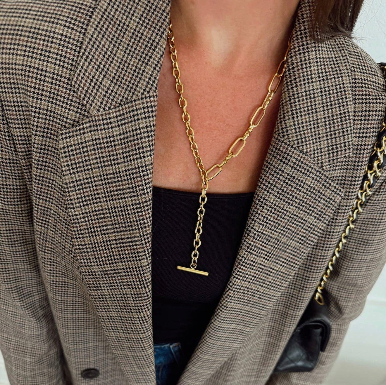 Person wearing a gold chain necklace with a t-bar clasp, partially visible outfit with a checkered jacket.