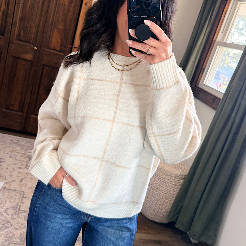 A woman takes a mirror selfie wearing a cream-colored, oversized knit sweater with a soft beige windowpane grid pattern and blue jeans.