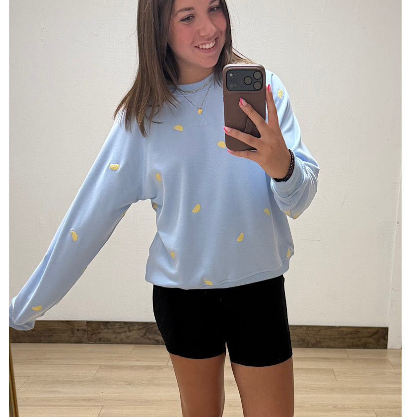 Person wearing a light blue sweatshirt with yellow patterns and black shorts, taking a mirror selfie.