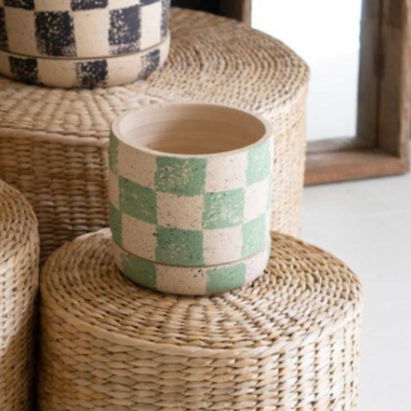 Three checkered ceramic plant pots in yellow, black-and-white, and mint green, displayed on a tiered stack of woven seagrass poufs with potted plants in a styled interior setting.