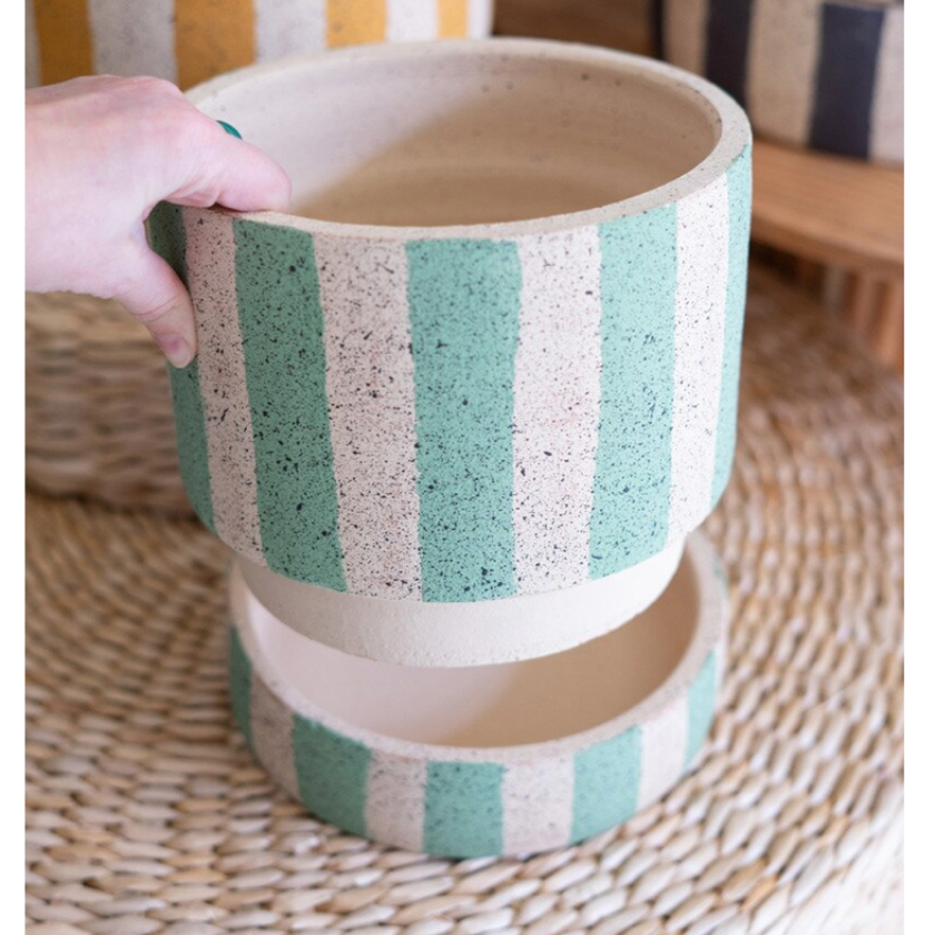 A hand holds a round planter pot with vertical mint green and cream speckled stripes, with a second matching pot stacked underneath on a woven mat.