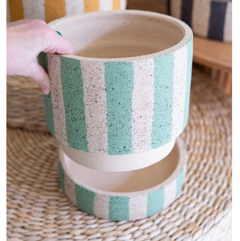 A hand holds a round planter pot with vertical mint green and cream speckled stripes, with a second matching pot stacked underneath on a woven mat.