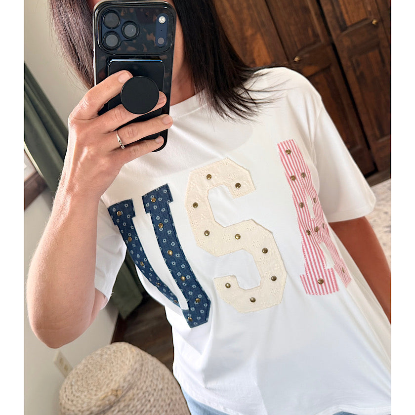 Person wearing a white t-shirt with decorative letters, taking a mirror selfie.