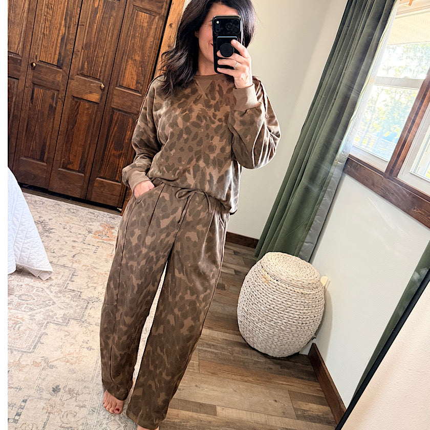 A woman takes a mirror selfie in a bedroom wearing a matching brown animal-print lounge set consisting of a long-sleeve top and wide-leg pants.