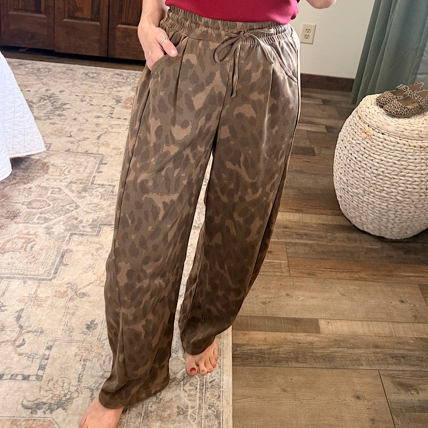 A person wearing loose, wide-leg drawstring pants in a brown and taupe leopard-like pattern, standing barefoot on a patterned rug in a home interior.