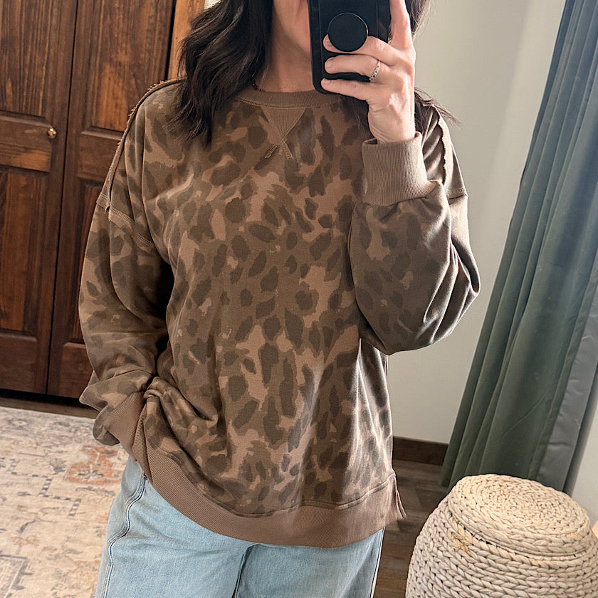 A woman takes a mirror selfie wearing a brown and tan leopard-print crewneck sweatshirt and light blue jeans in a home setting.