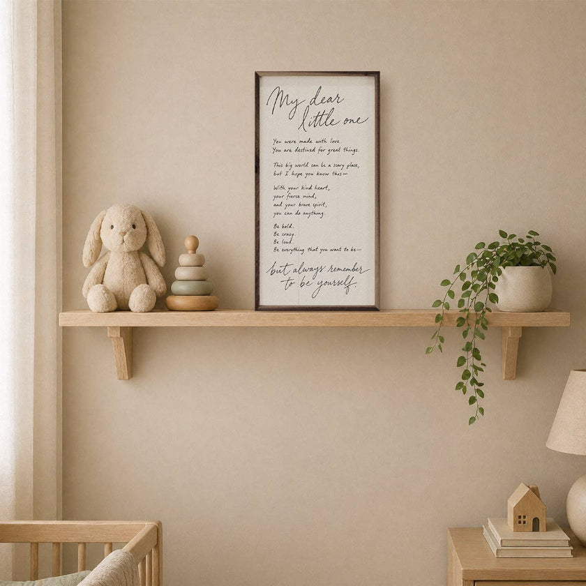 A framed wall art print with a poem titled 'My dear little one' displayed on a wooden shelf in a nursery setting, alongside a plush bunny, stacked wooden toys, a potted plant, and a crib.