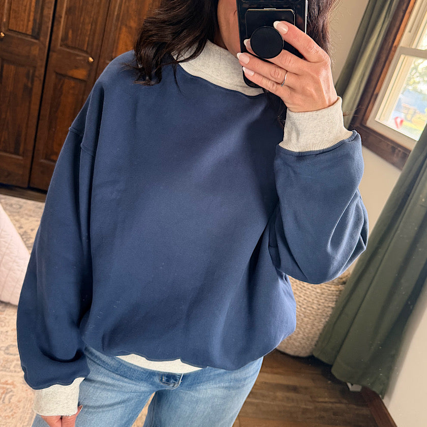 A person taking a mirror selfie in a home setting, wearing an oversized navy blue sweatshirt with a light gray mock neck collar and ribbed cuffs, paired with light blue jeans.