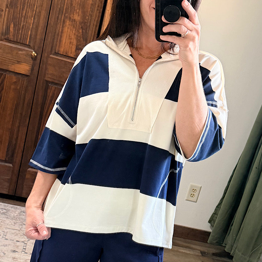 A woman takes a mirror selfie wearing a color-blocked quarter-zip pullover top with navy blue and cream horizontal stripes, paired with navy pants in a home setting.