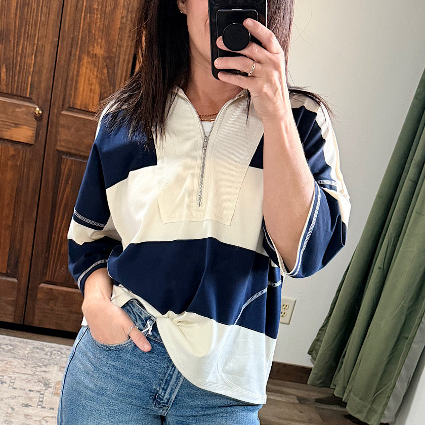A woman takes a mirror selfie wearing a navy blue and cream color-blocked quarter-zip pullover top with blue jeans, set in a home environment with a wooden door and green curtain in the background.