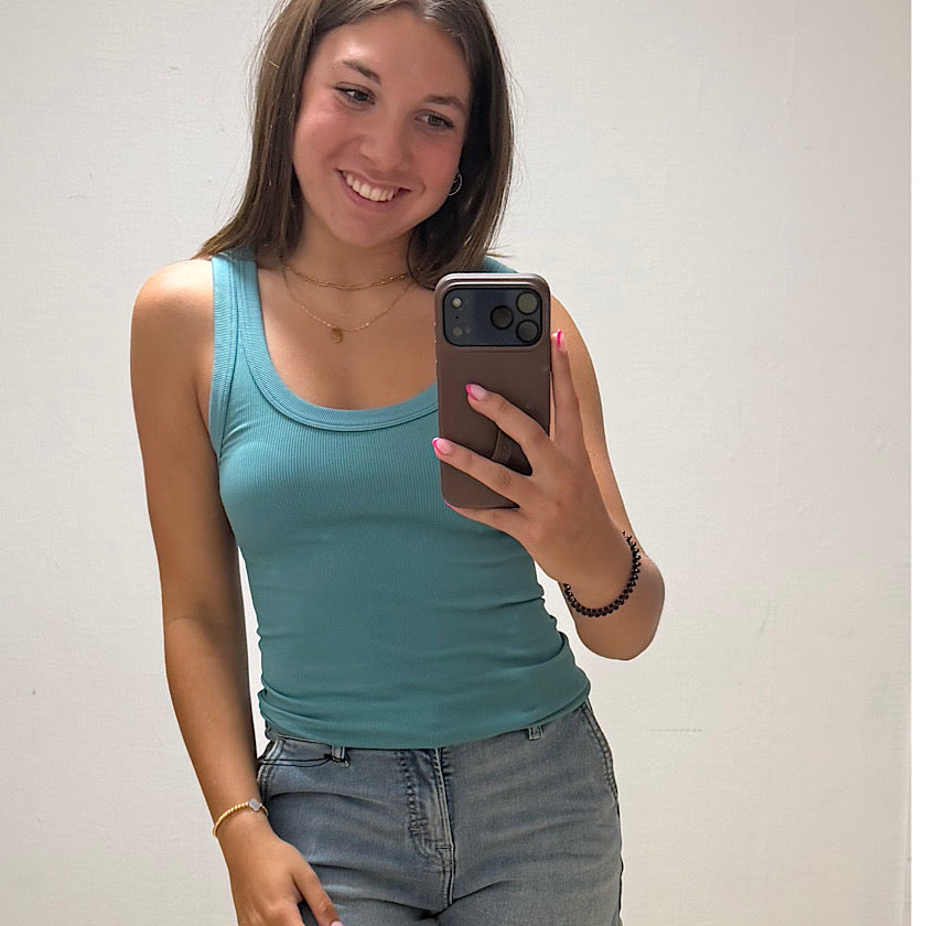 Woman taking a mirror selfie wearing a teal tank top and jeans.