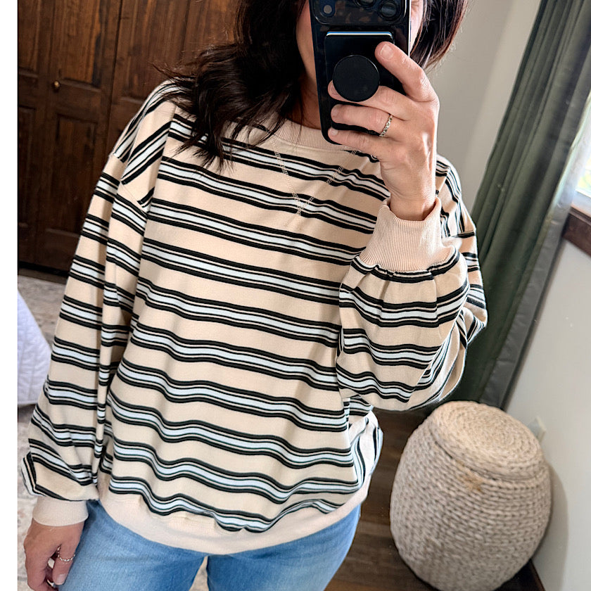 A woman takes a mirror selfie wearing a cream-colored crewneck sweatshirt with thin black horizontal stripes, paired with blue jeans in a bedroom setting.