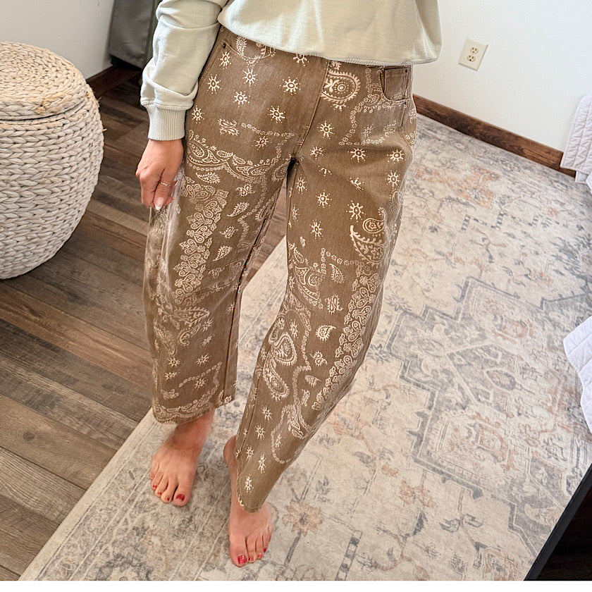 A person wearing brown pants with a beige paisley and floral pattern, standing barefoot on a patterned area rug in a room.
