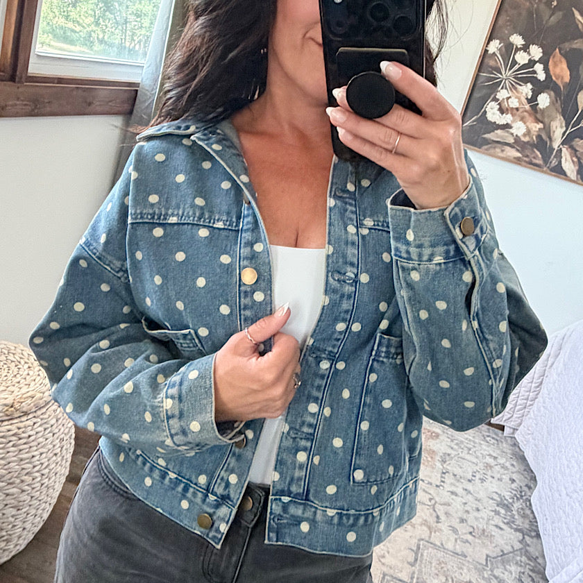 A woman takes a mirror selfie wearing a cropped blue denim jacket with a white polka dot pattern over a white tank top.