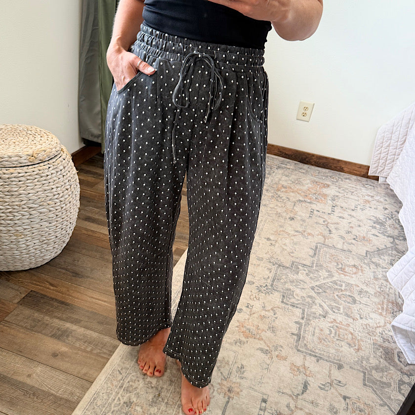 A person wearing loose black drawstring pants with small white polka dots, standing barefoot on a patterned rug in a bedroom setting.