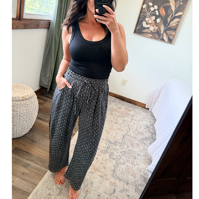 A woman takes a mirror selfie in a bedroom while wearing a black ribbed tank top and dark gray wide-leg pants with small white polka dots and a drawstring waist.