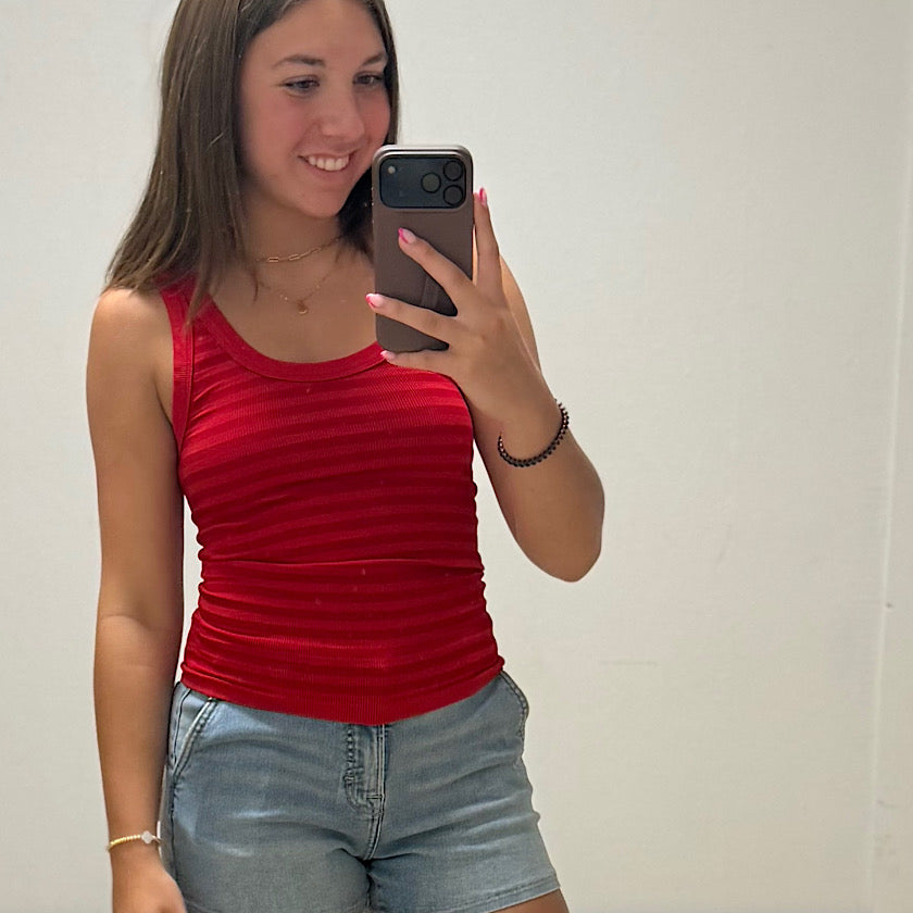 Woman taking a mirror selfie wearing a red tank top and denim shorts.