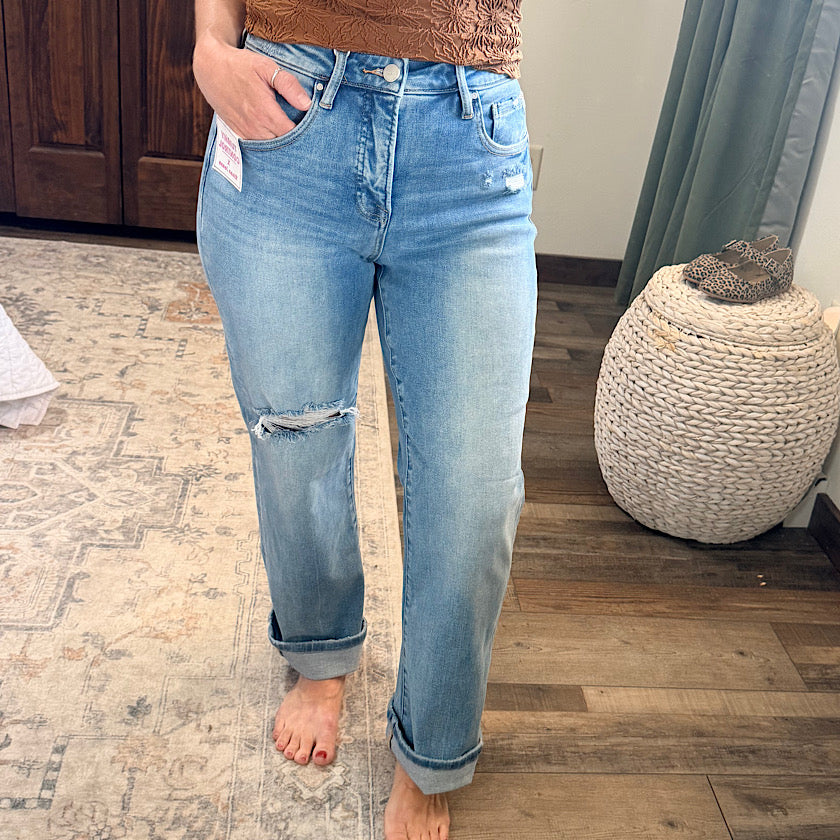 A person wearing light-wash blue distressed jeans with a ripped knee detail and cuffed hems, standing barefoot on a patterned rug in a home setting.