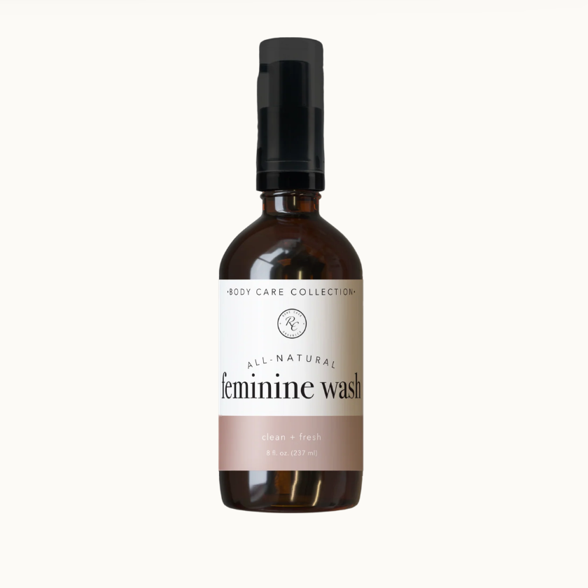 A bottle of Feminine Wash body care product from the Body Care Collection, featuring a dark amber glass bottle, a black cap, and a white label with a dusty pink band at the bottom.