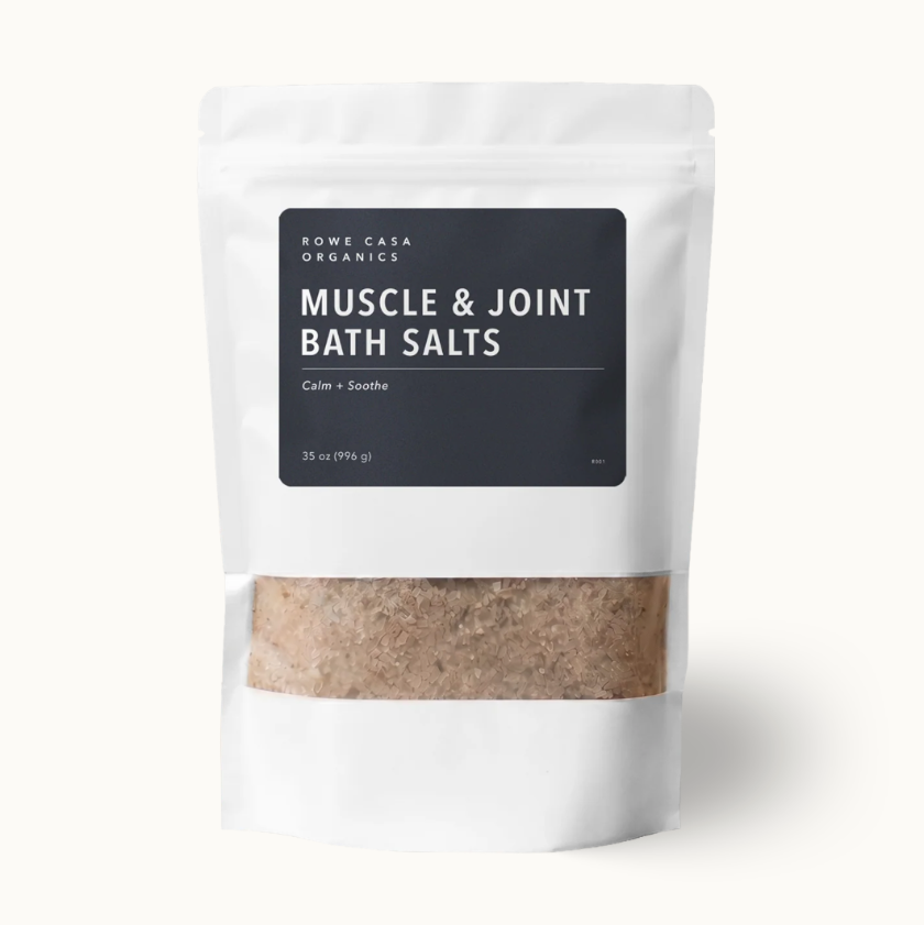 A white stand-up pouch of Rowe Casa Organics Muscle & Joint Bath Salts, featuring a black label and a clear window showing the beige-brown bath salts inside, set against a clean off-white background.